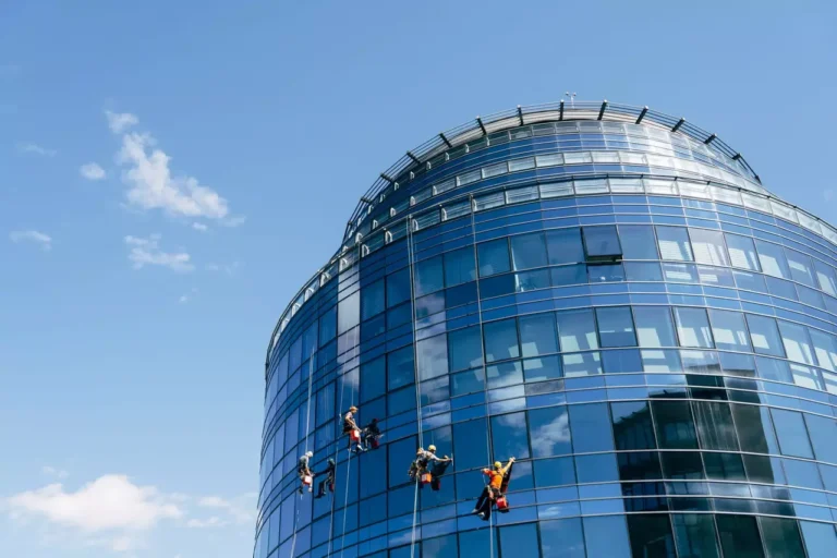 Dieses Bild zeigt ein modernes, hoch aufragendes Bürogebäude mit einer gebogenen Glasfassade. Mehrere Arbeiter, die Sicherheitsausrüstung tragen, sind an Seilen befestigt und reinigen die Glasflächen des Gebäudes. Der Himmel ist klar und blau, was eine helle und klare Umgebung für die Arbeit schafft. Die Szene zeigt deutlich die Höhe des Gebäudes und die Herausforderungen der Fassadenreinigung.